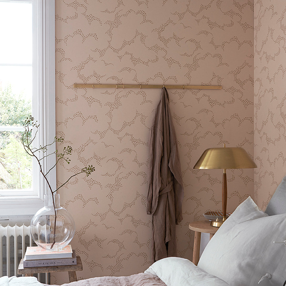 Beige patterned bedroom corner with wooden coat rack, brown robe, brass lamp, wood side table, glass vase, and gray bedding.