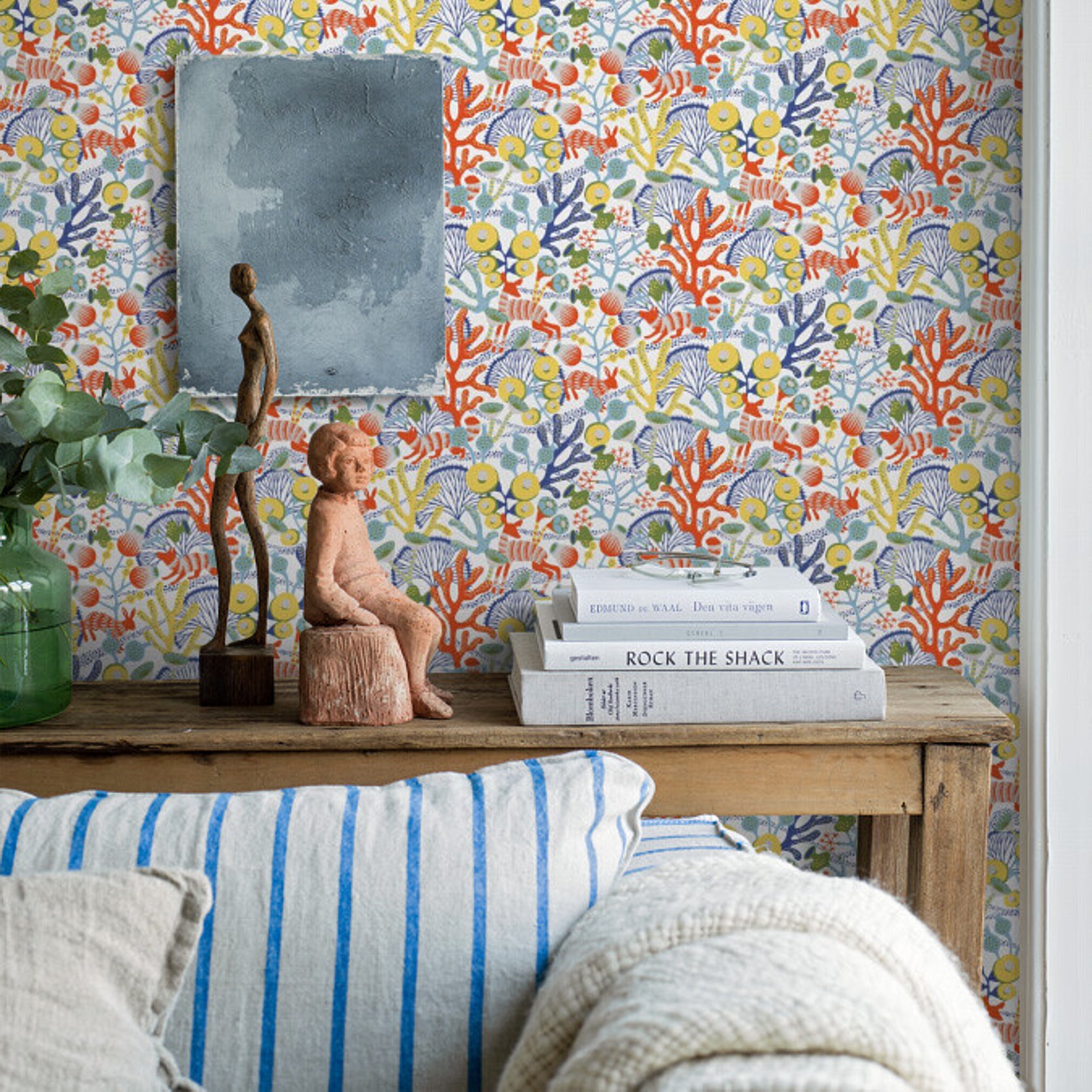 Rustic wooden console with coral-pattern wallpaper, slate plaque, figurines, white books, green plant, blue-striped cushion.