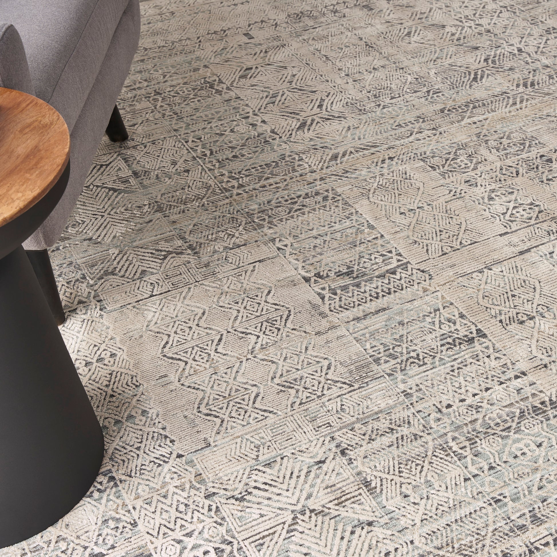 Geometric beige-gray patterned rug with diamond/chevron motifs; partial view of a gray sofa and round wooden table.