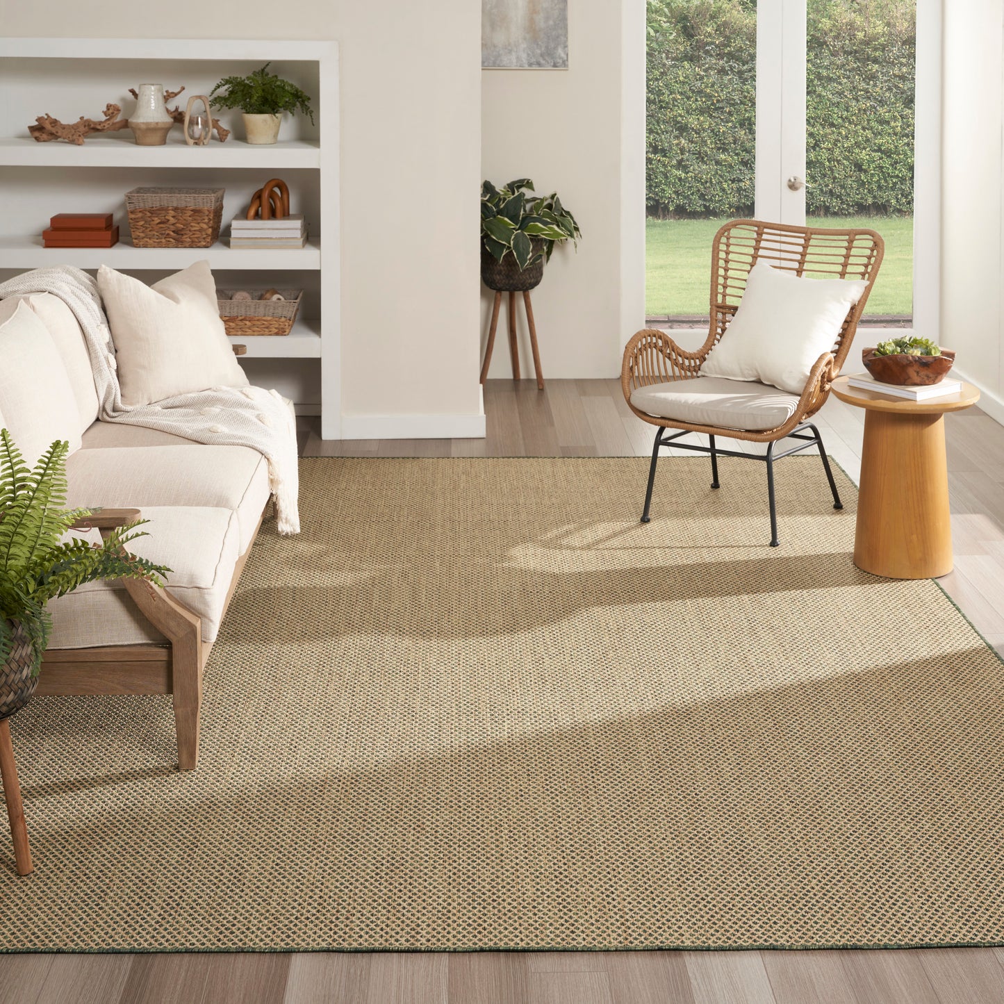 Neutral living room with a beige woven rug, cream sofa, rattan chair, wooden side table, plants, and glass doors.