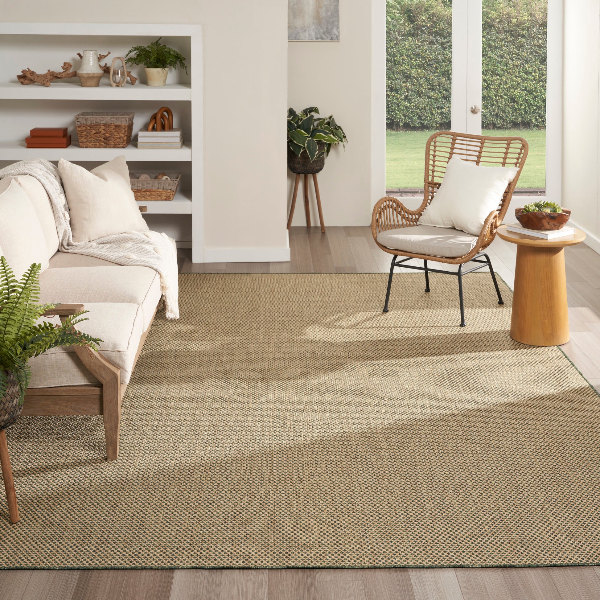 Neutral living room with a beige woven rug, cream sofa, rattan chair, wooden side table, plants, and glass doors.