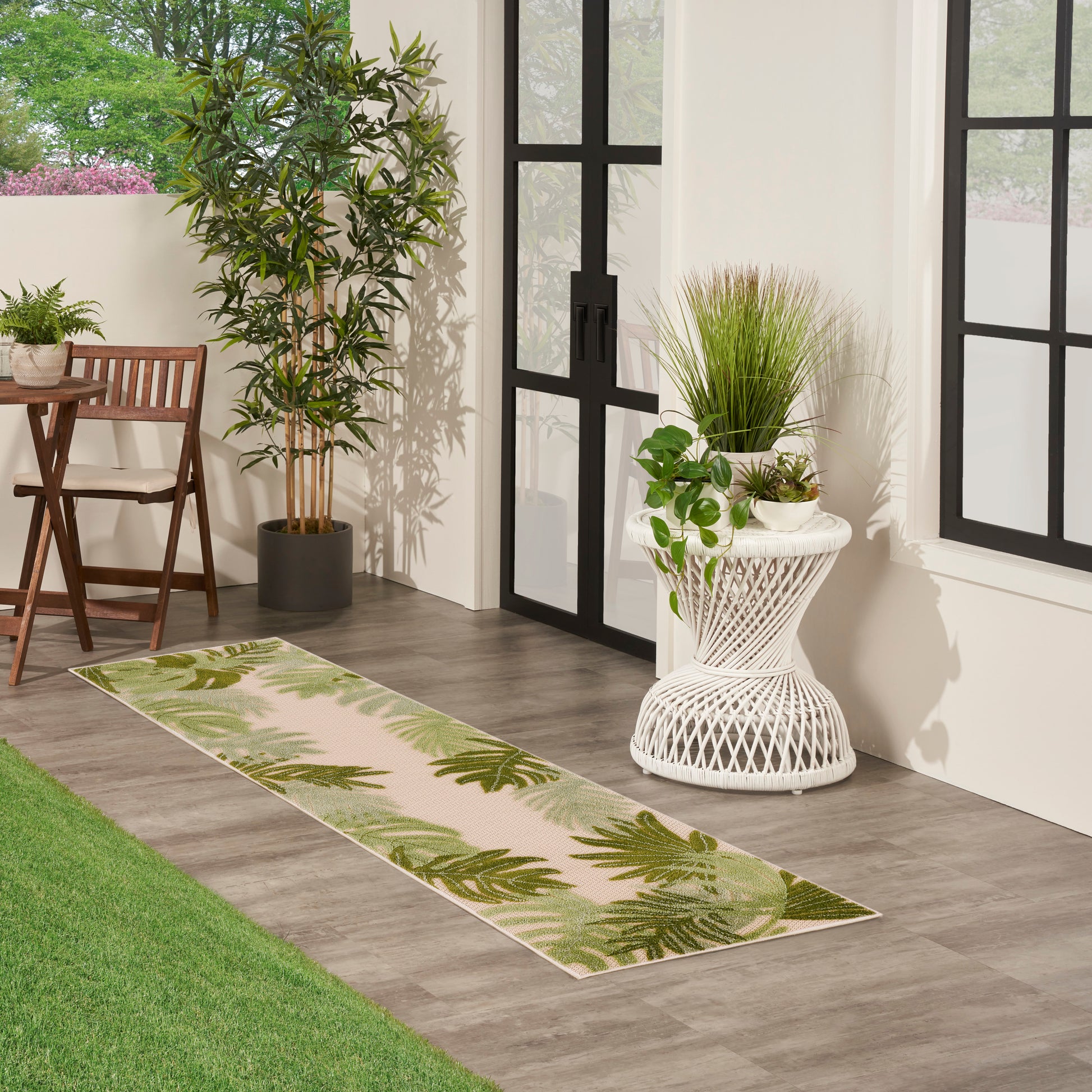 Beige-green palm leaf runner rug on gray tile beside potted plants and white wicker stand.