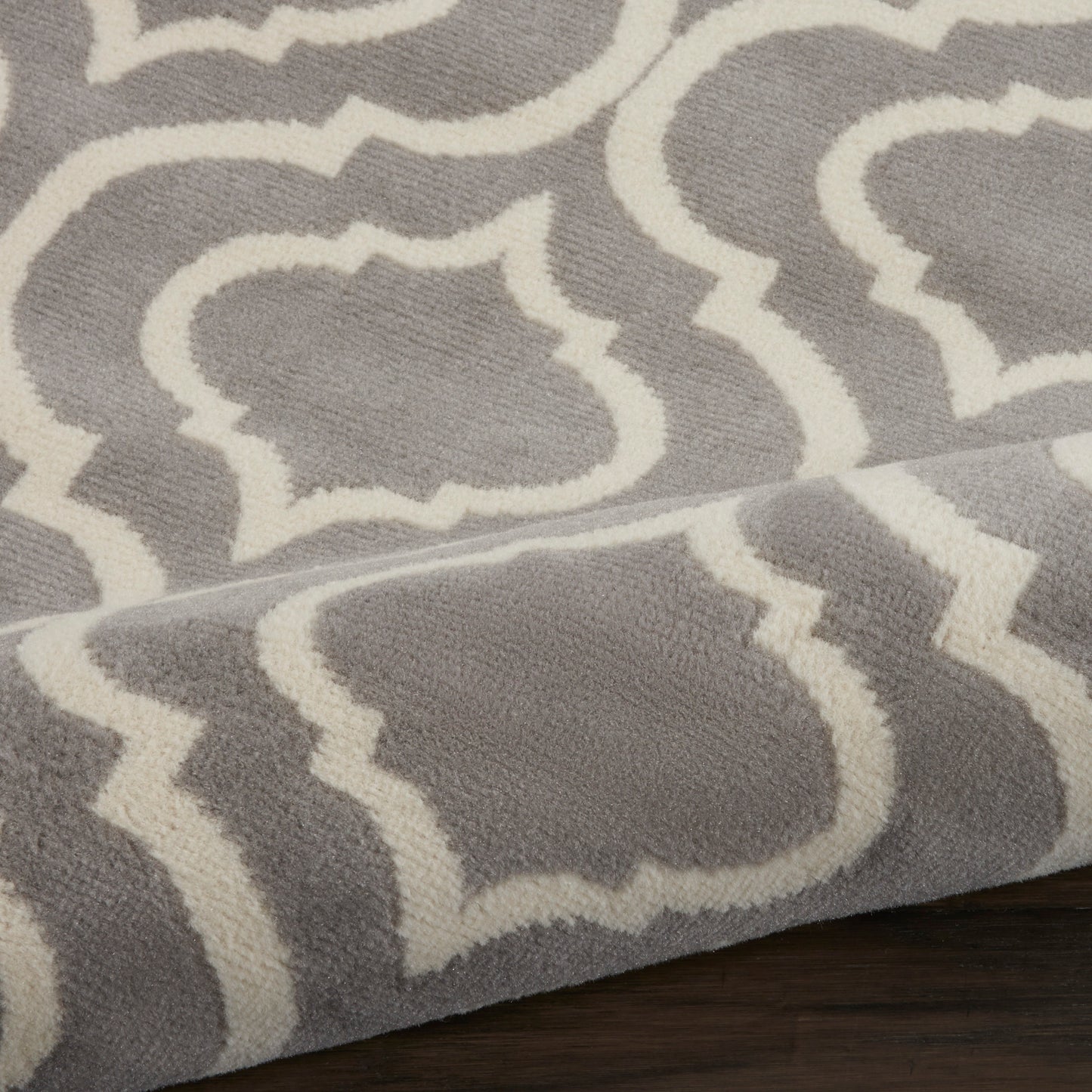Gray plush rug with white trellis/quatrefoil pattern; soft pile, folded edge on dark wood floor.
