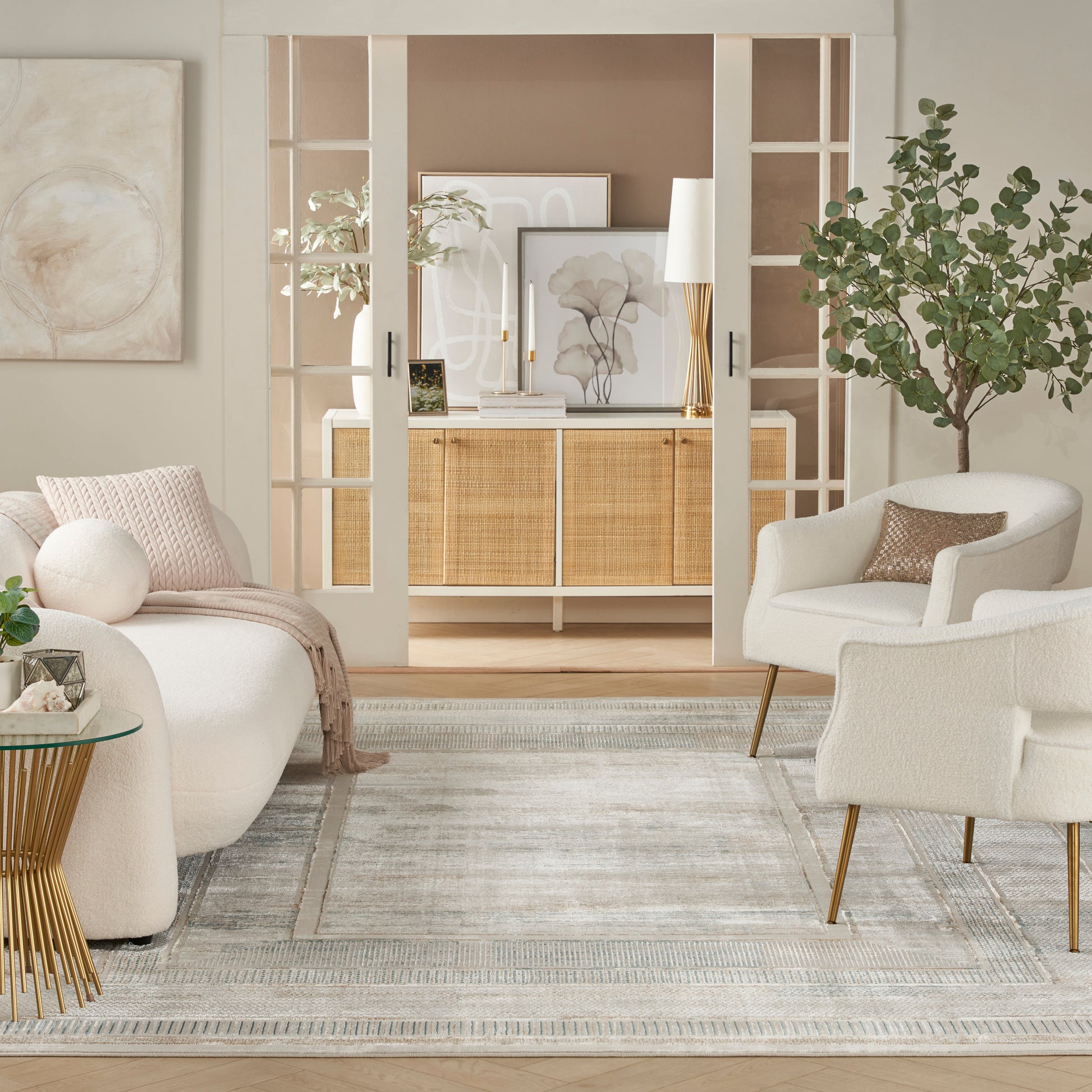Bright, cozy living room with white sofas and armchairs, cane cabinet, glass doors, greenery, beige area rug.