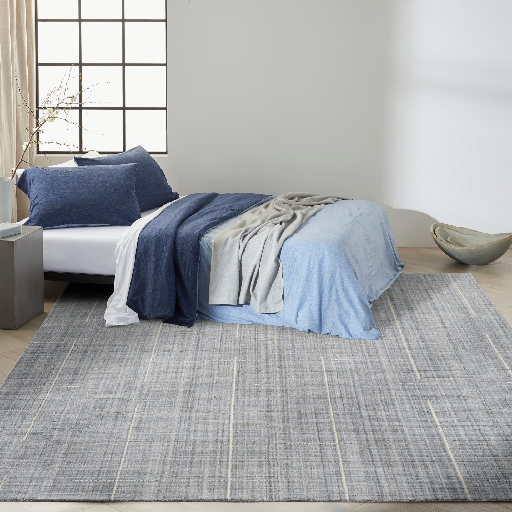 Modern bedroom with a bed dressed in navy, light blue and gray linens, layered throws; grey striped rug on light wood floor.