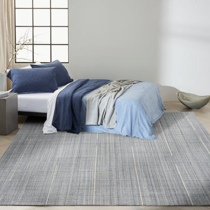 Modern bedroom with a bed dressed in navy, light blue and gray linens, layered throws; grey striped rug on light wood floor.
