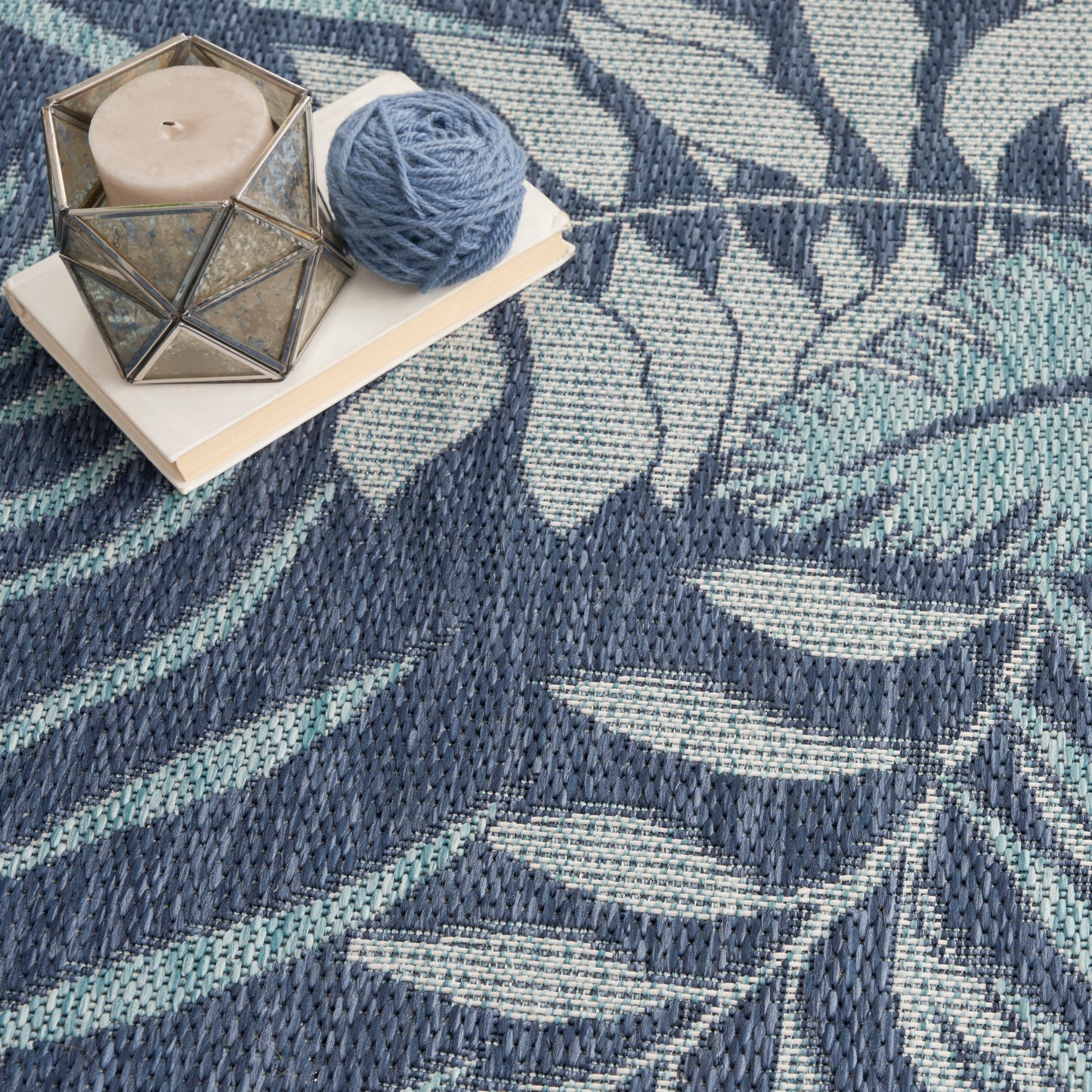 Round blue leafy woven outdoor rug with a geometric glass candle holder, beige candle, blue yarn ball, and white notebook on it.