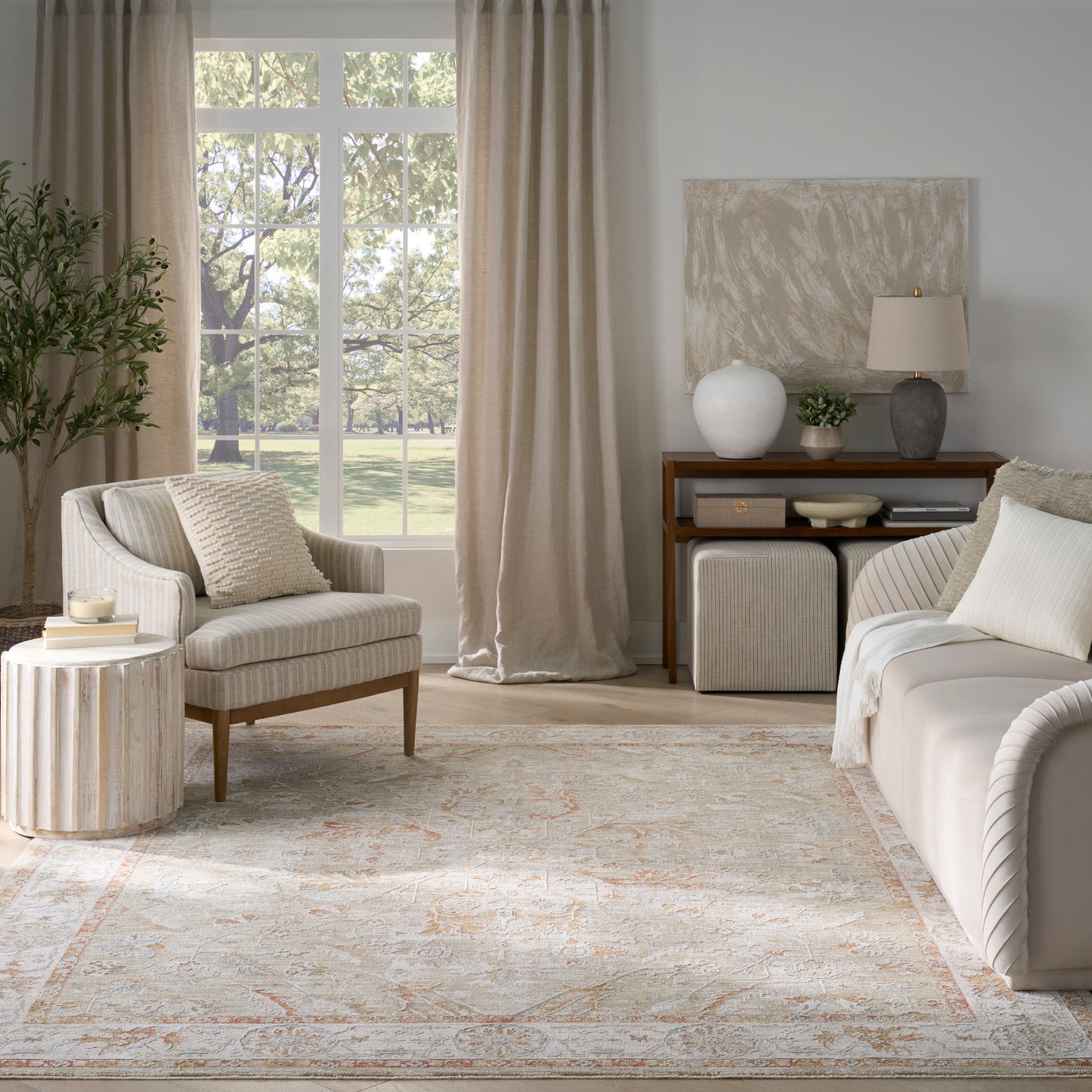 Neutral living room with beige curtains, cream upholstered chair, round wood side table, pale patterned rug, white sofa and wooden console with lamps.