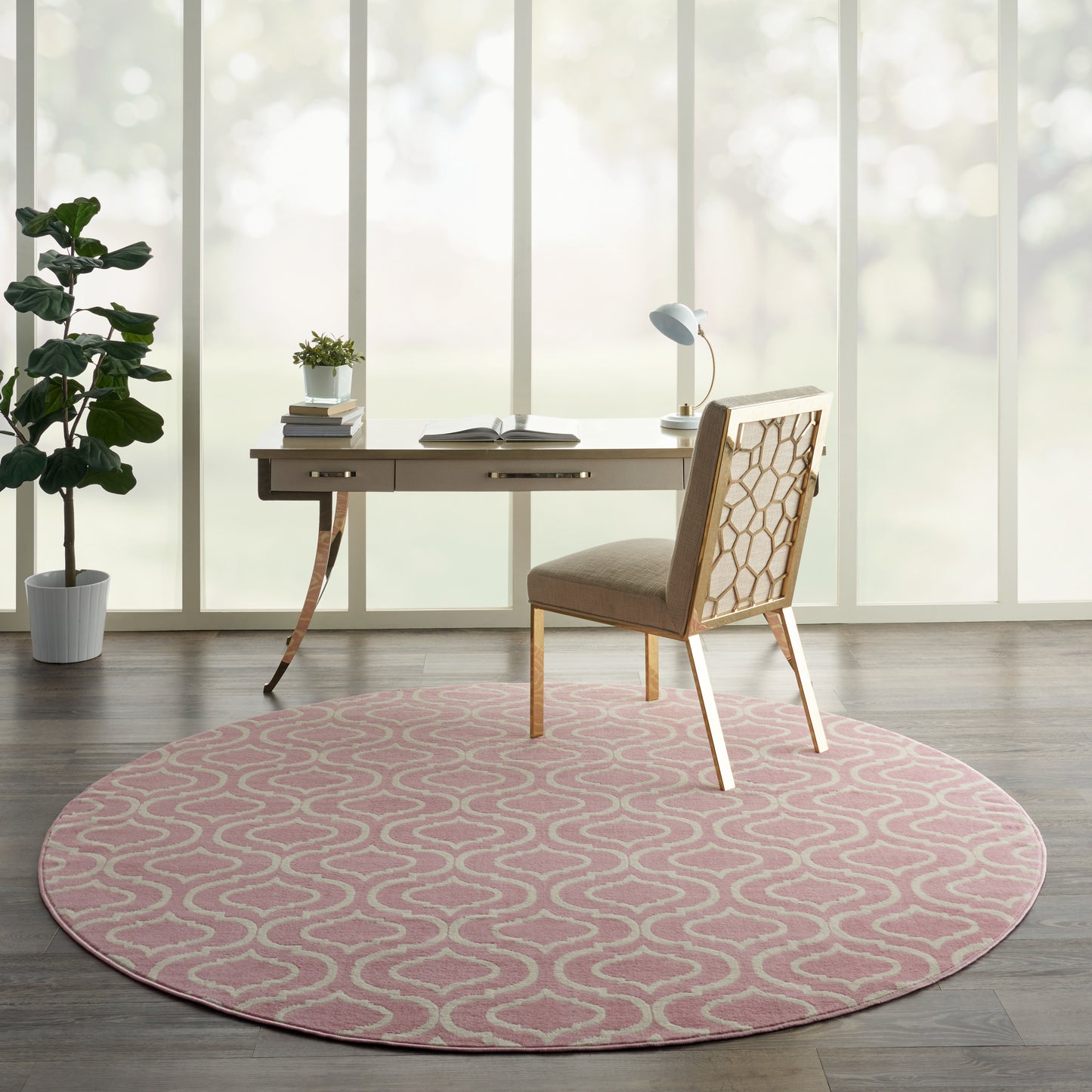 Round pink rug with white geometric pattern under a wooden desk and beige upholstered chair in a sunlit home office.