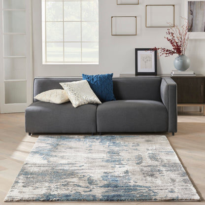 Abstract blue-gray rug on light wood floor with a side table and vase visible.