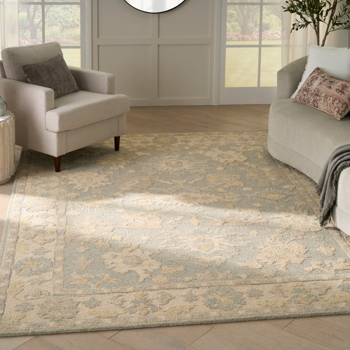 Neutral living room with a beige ornate area rug, beige armchair, curved gray sofa, round mirror, and large windows.