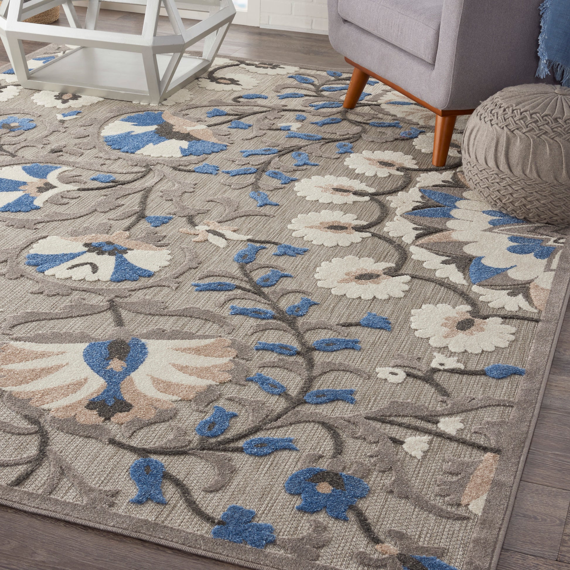 Beige floral area rug with blue and ivory blooms and gray vines, in a modern living room with a gray chair and knitted pouf.