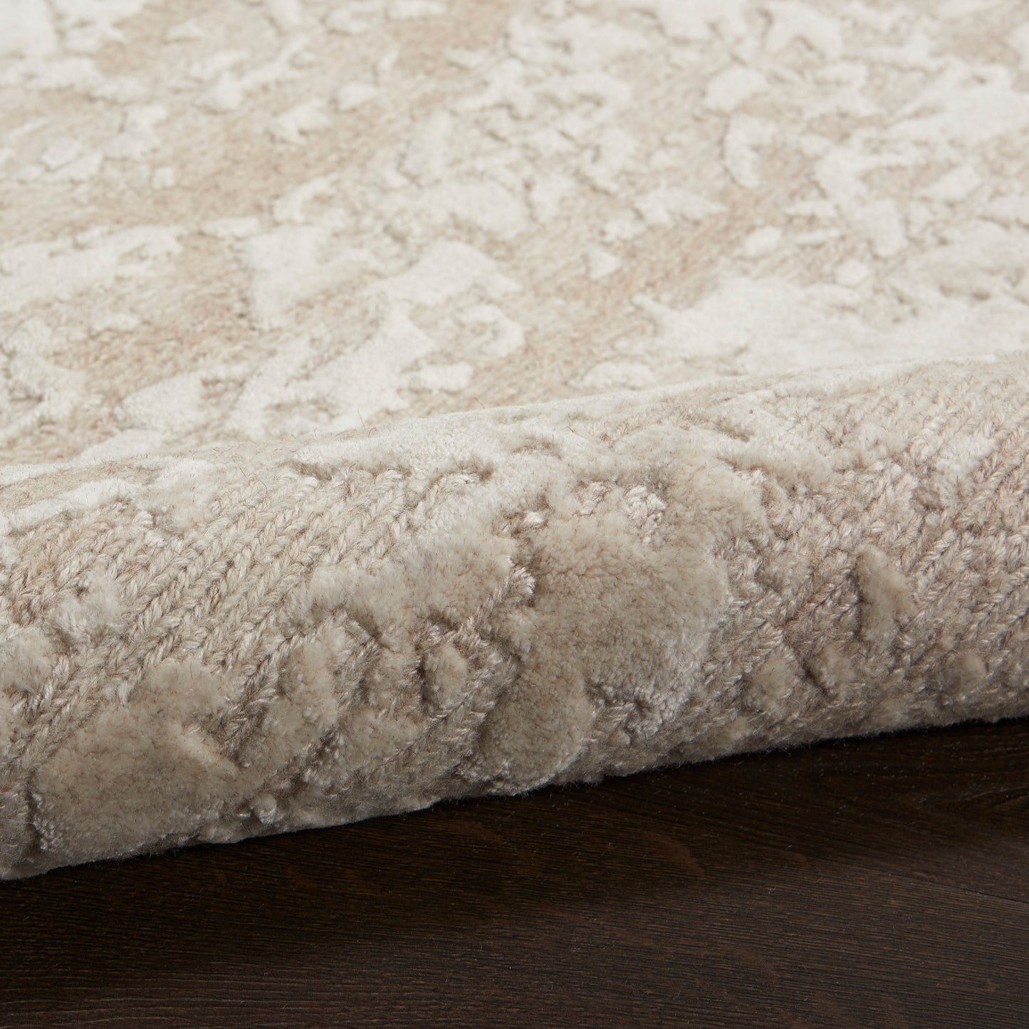 Beige patterned plush area rug with soft pile and rolled edge on dark wood floor.