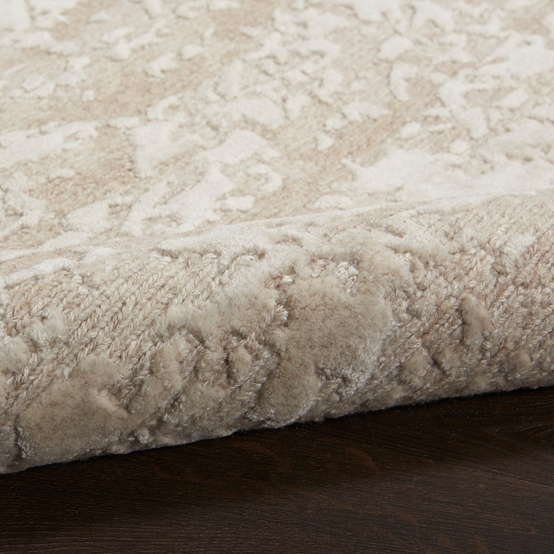 Beige patterned plush area rug with soft pile and rolled edge on dark wood floor.