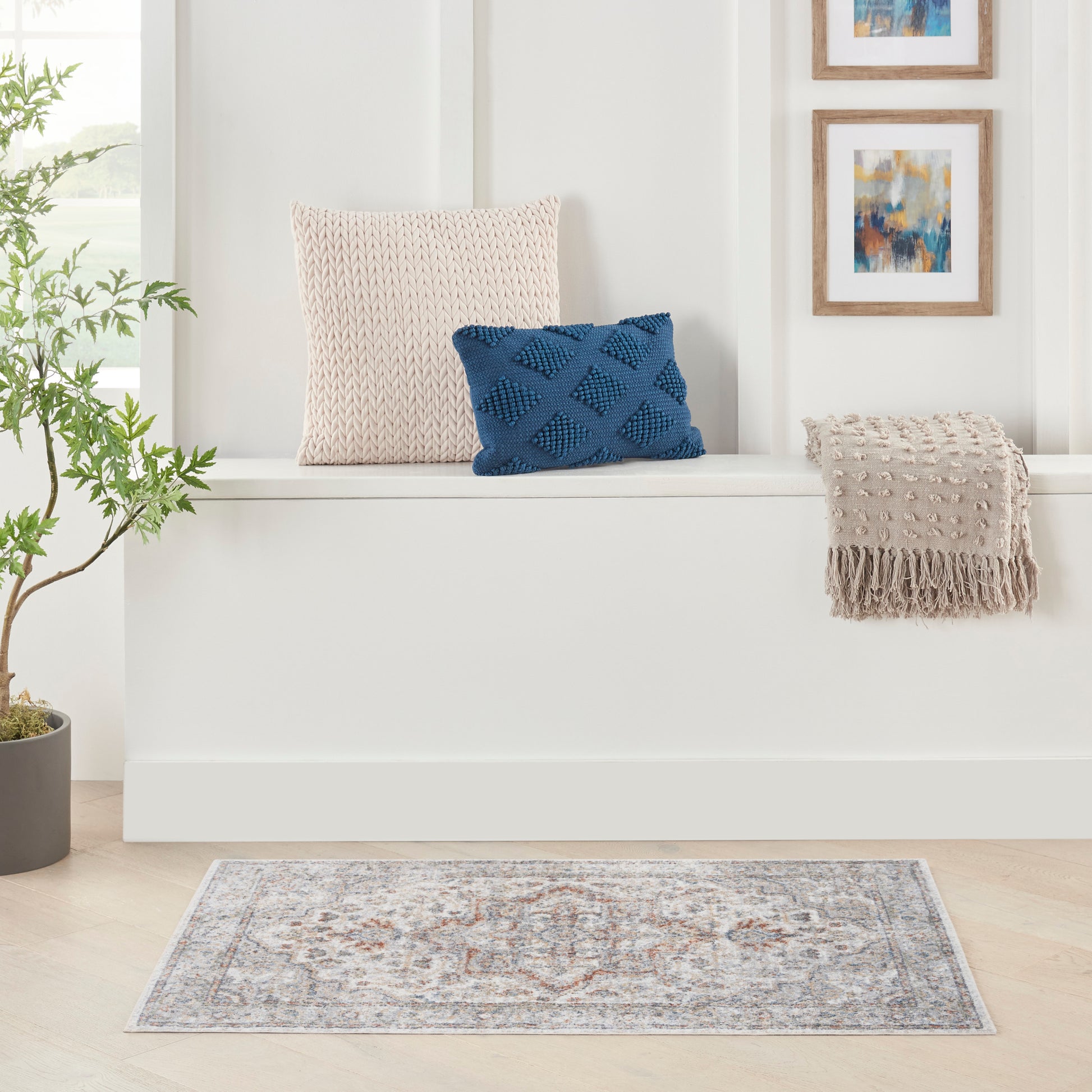 Cream knitted cushion, blue textured pillow, beige fringed throw on a white bench with plant, art, and a patterned rug.