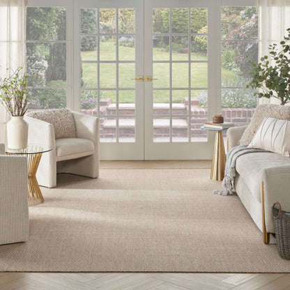 Bright living room with beige sofas and armchairs, a woven rug, glass French doors, brass accents, and greenery outside