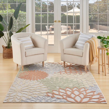 Two beige fabric armchairs with striped pillows and a draped throw blanket sit on a pastel leaf rug by sunlit glass doors.