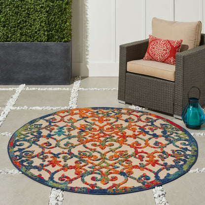 Round multicolor outdoor rug with an intricate scroll pattern beside a beige cushioned wicker chair on a stone patio.