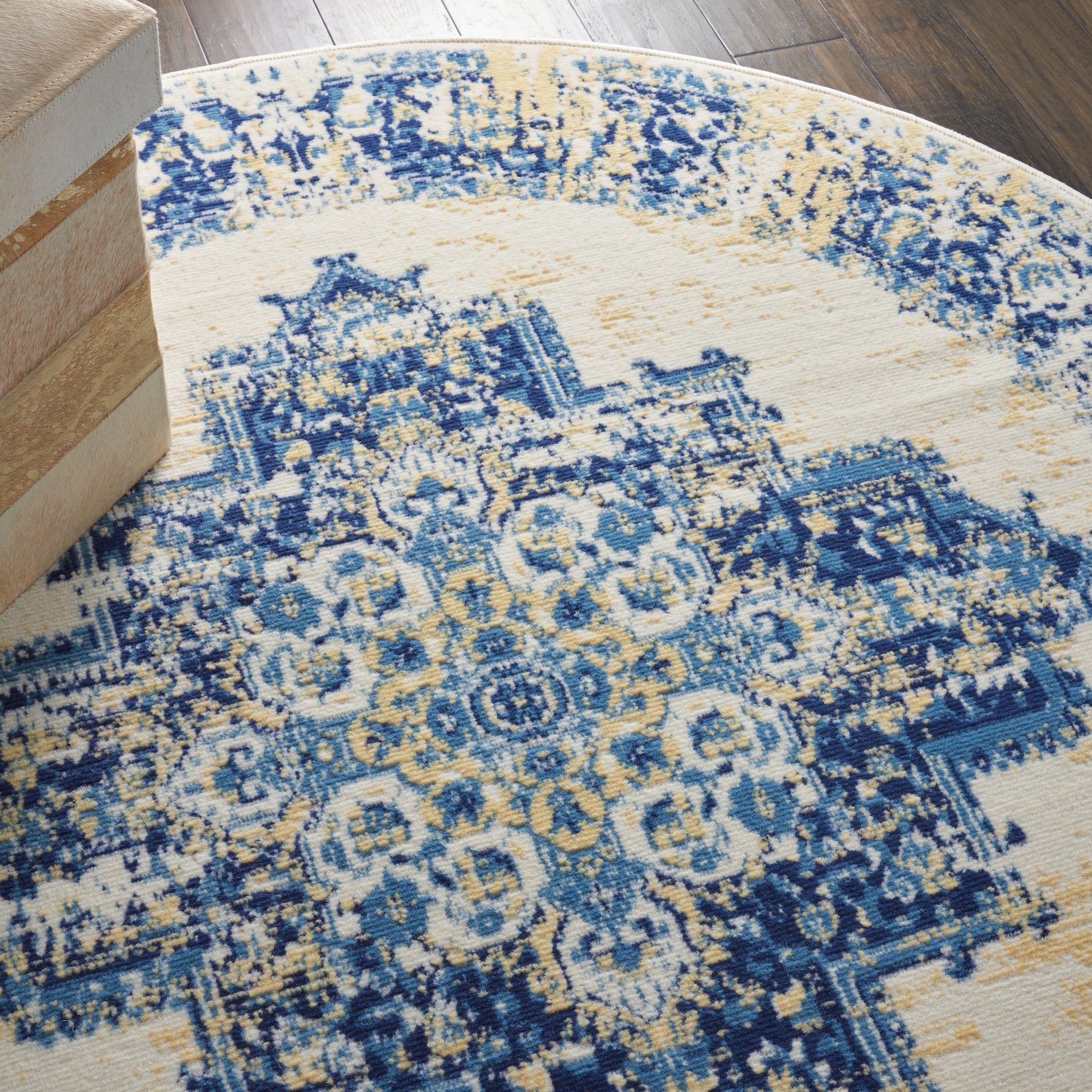 Round rug with blue and yellow distressed oriental pattern on cream field, on dark wood floor near stacked boxes.