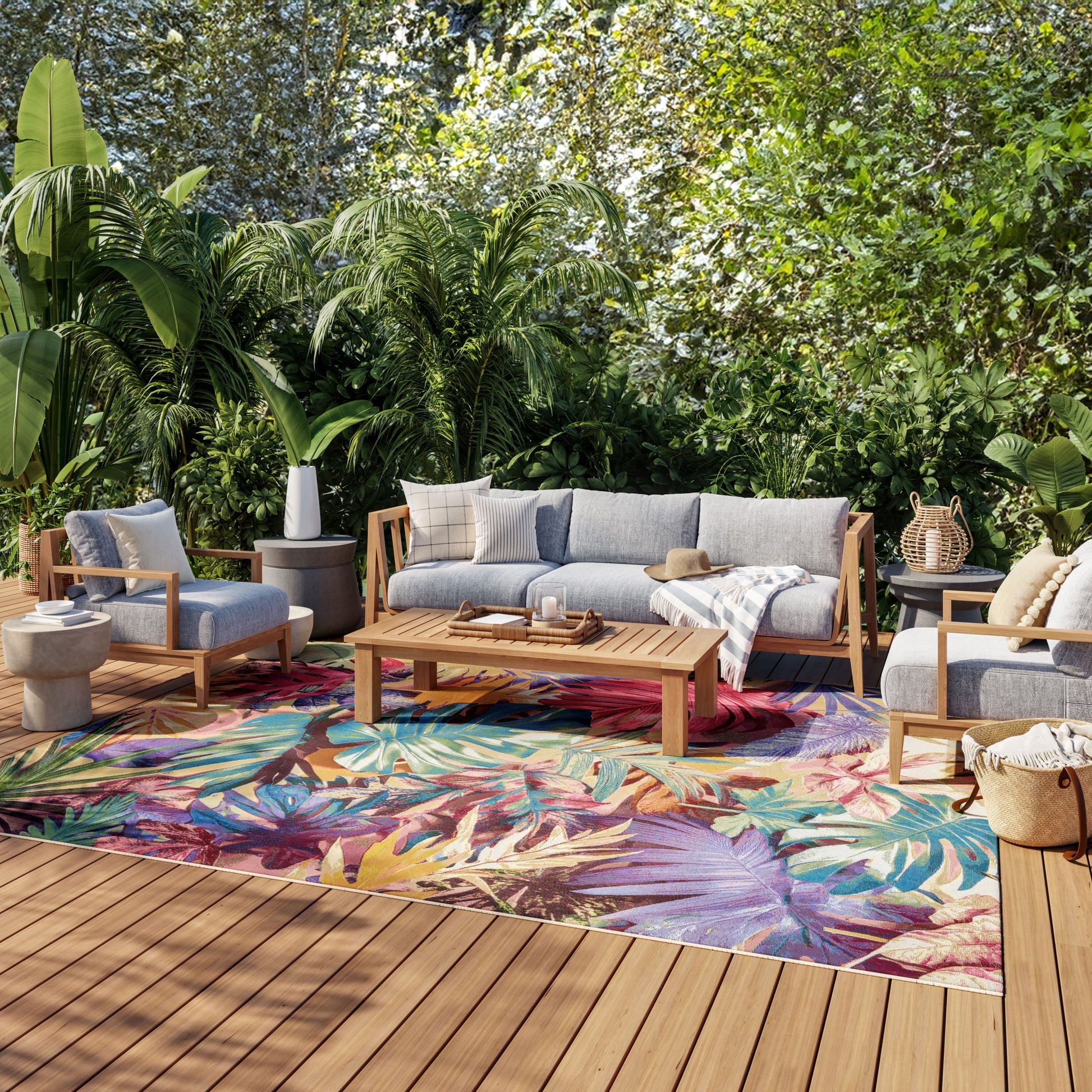 Outdoor lounge with gray cushioned wooden seating, vibrant tropical rug, coffee table, lanterns, and lush greenery.