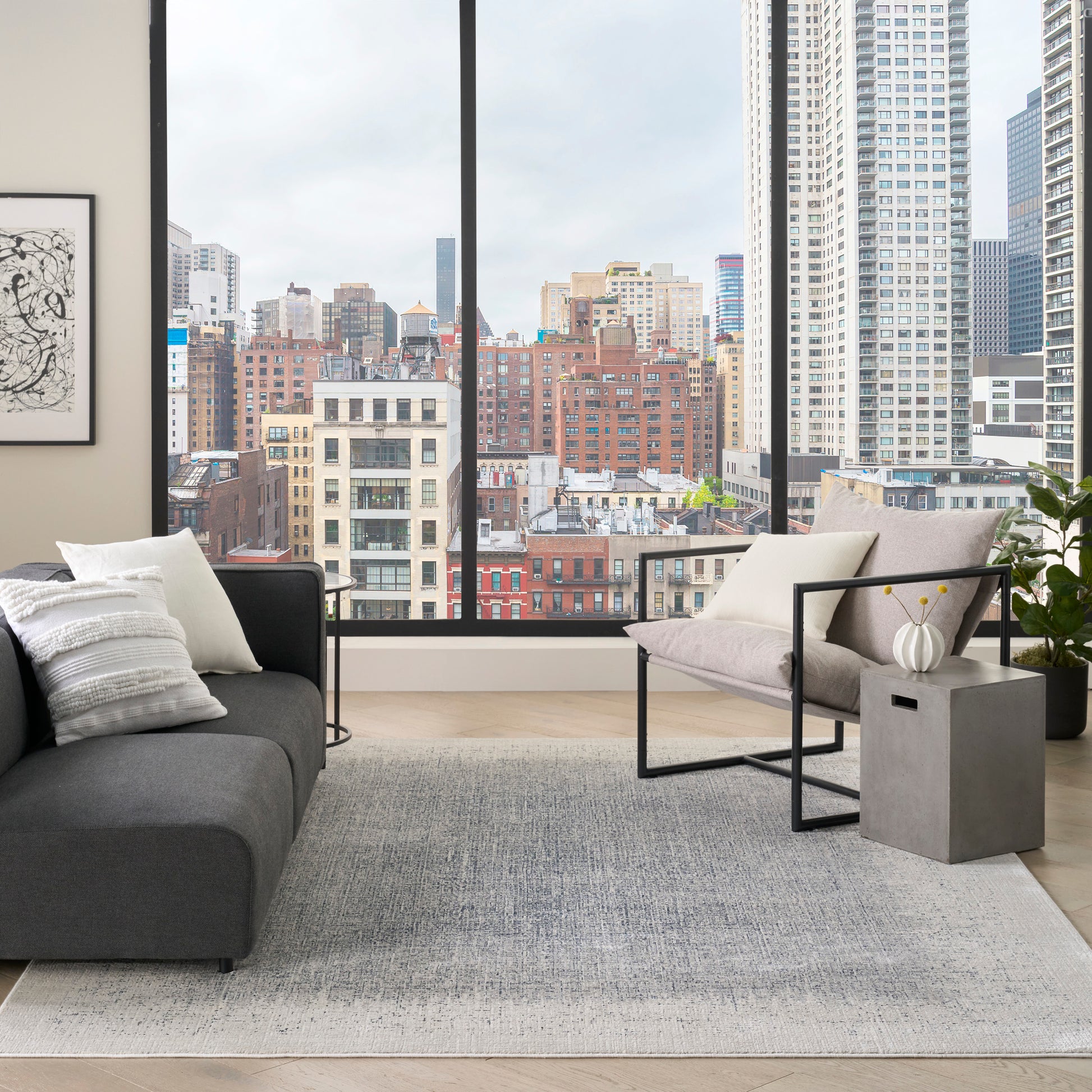 Modern living room with floor-to-ceiling windows, gray sectional, beige chair, metal frame chairs, rug, city skyline view.