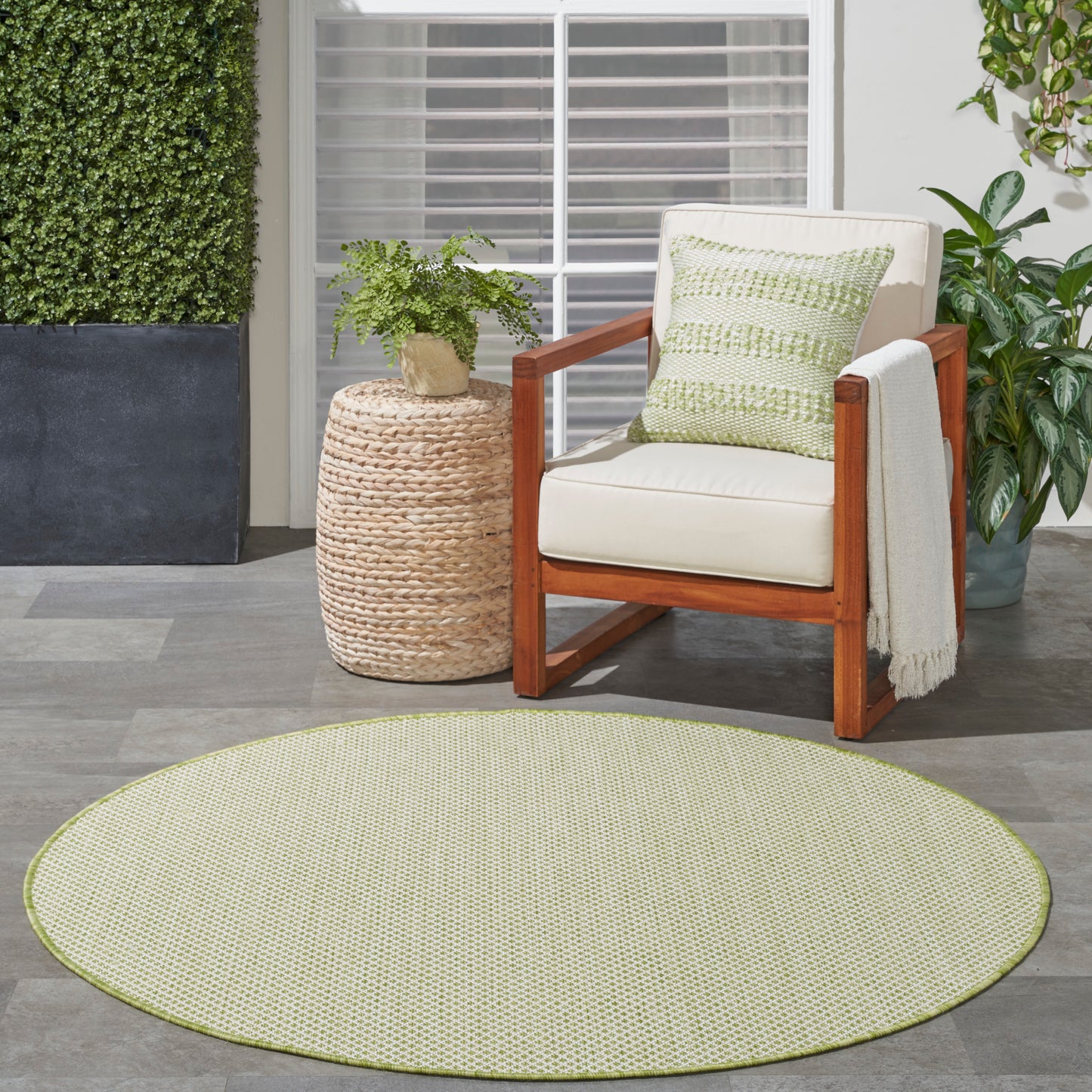 Round light-green textured rug, cream cushioned wooden armchair with green pillow and throw, woven side table, and plant.