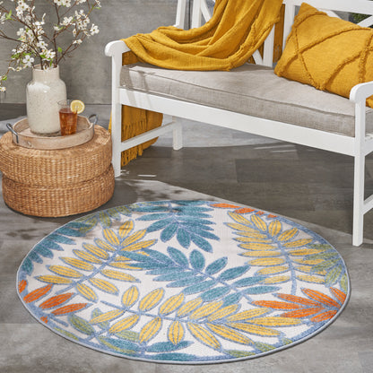 Round rug with blue, yellow and orange botanical leaves on a light gray background, beside a white bench with mustard throw.