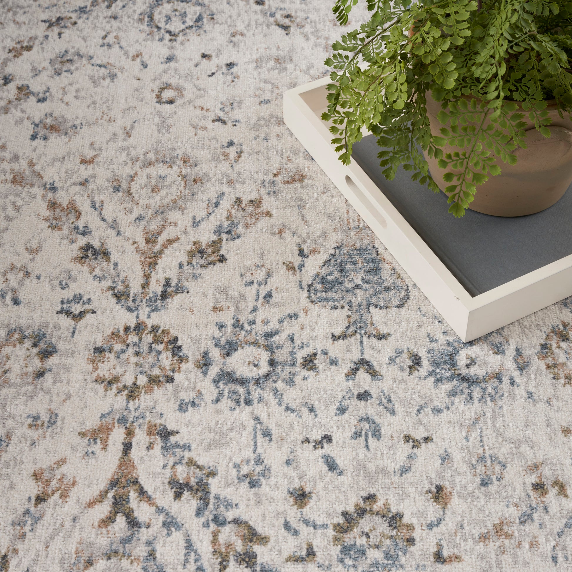 Light vintage floral area rug in blue, gray, beige; potted fern on a white tray atop it.
