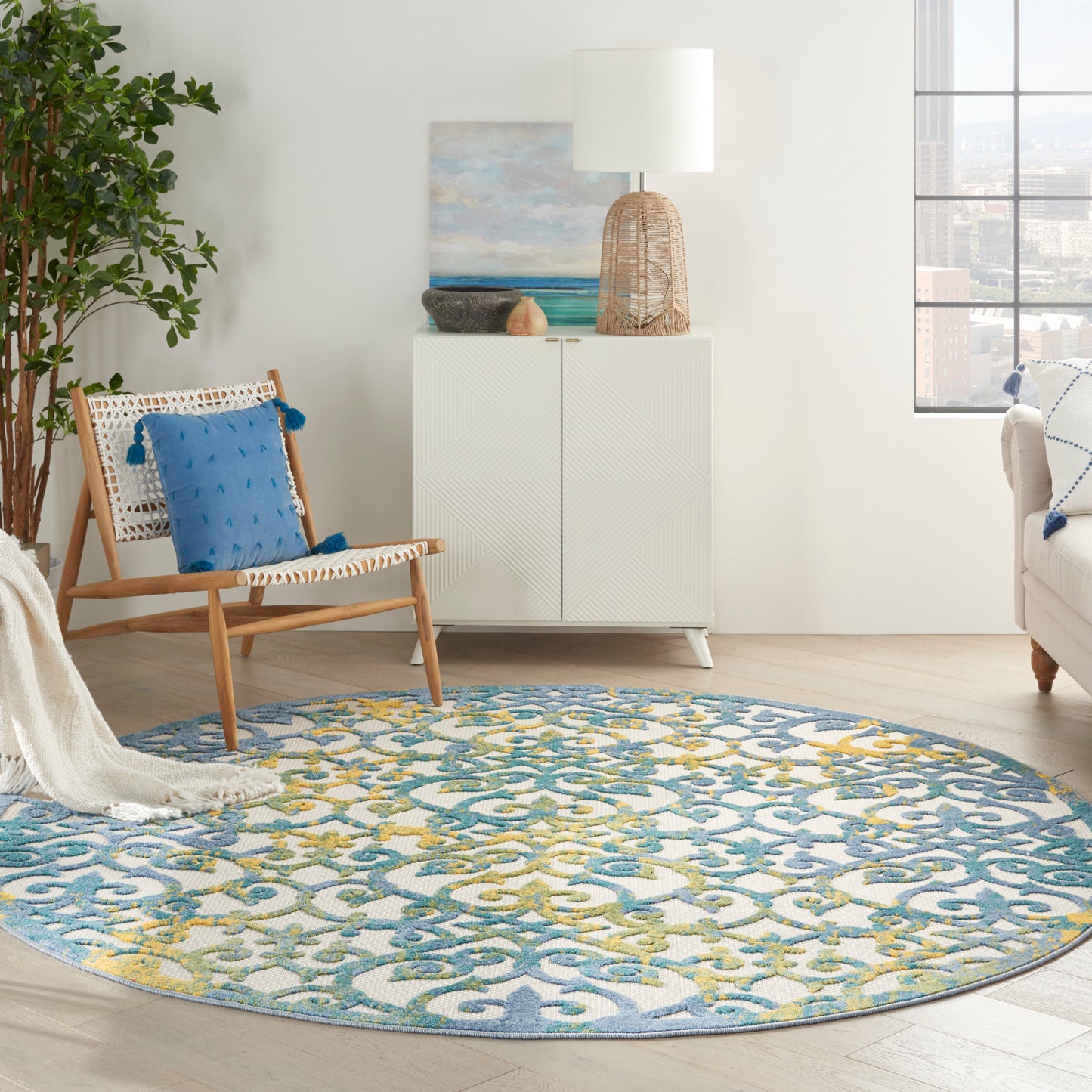 Bright living room with a round blue-yellow rug, woven chair with blue cushion, white cabinet, wicker lamp, plant, and view.