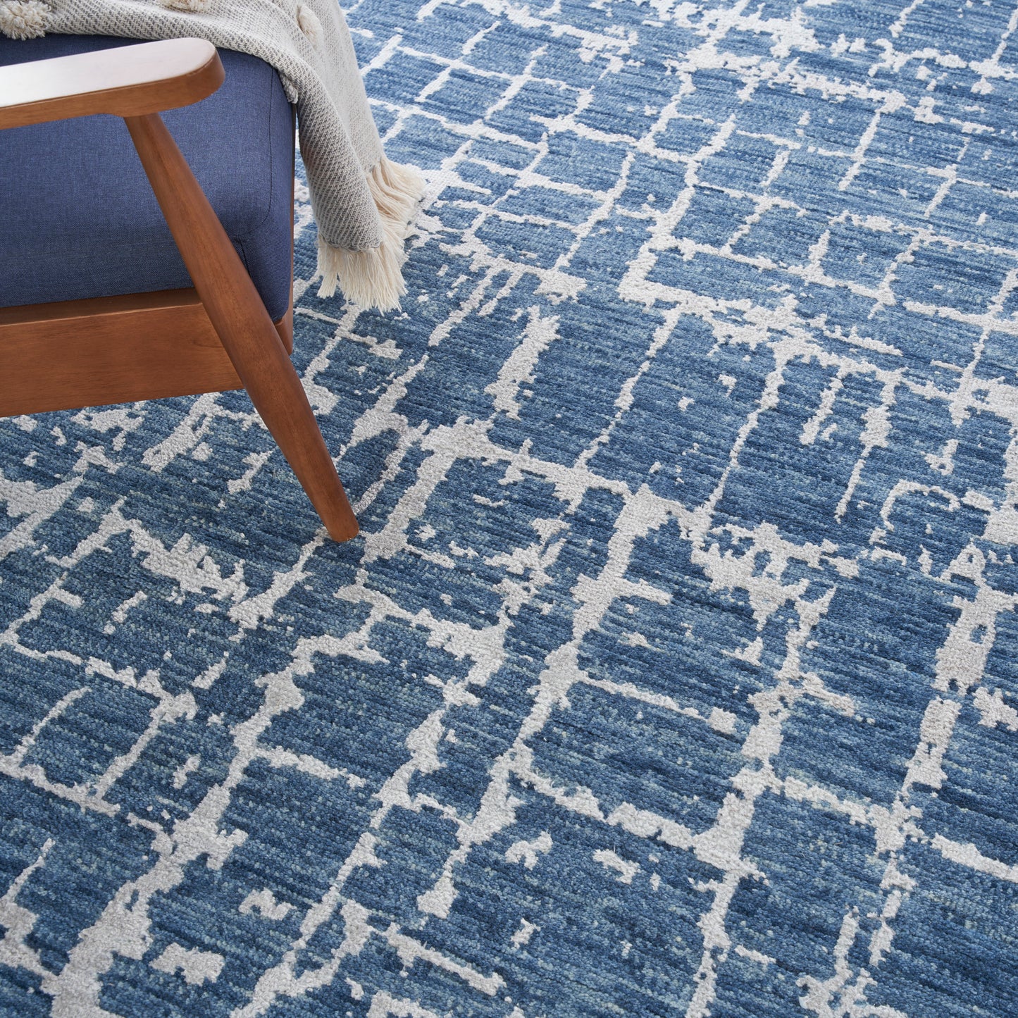 Blue geometric area rug with cream lattice pattern; wooden chair leg with blue upholstery and cream knitted throw.