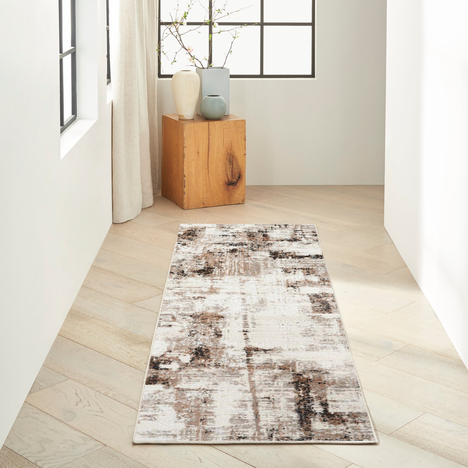 Neutral hallway with light wood floors, abstract brown-beige rug runner, and a wooden cube table with ceramic vases by a window.