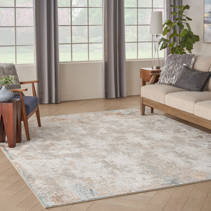Rectangular abstract area rug in beige, gray and blue tones on light wood floor, with beige sofa and blue chair.