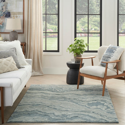 Bright living room with large black-framed windows, light gray sofa, wooden armchair, blue-gray wavy rug (5' x 7') & a potted plant.