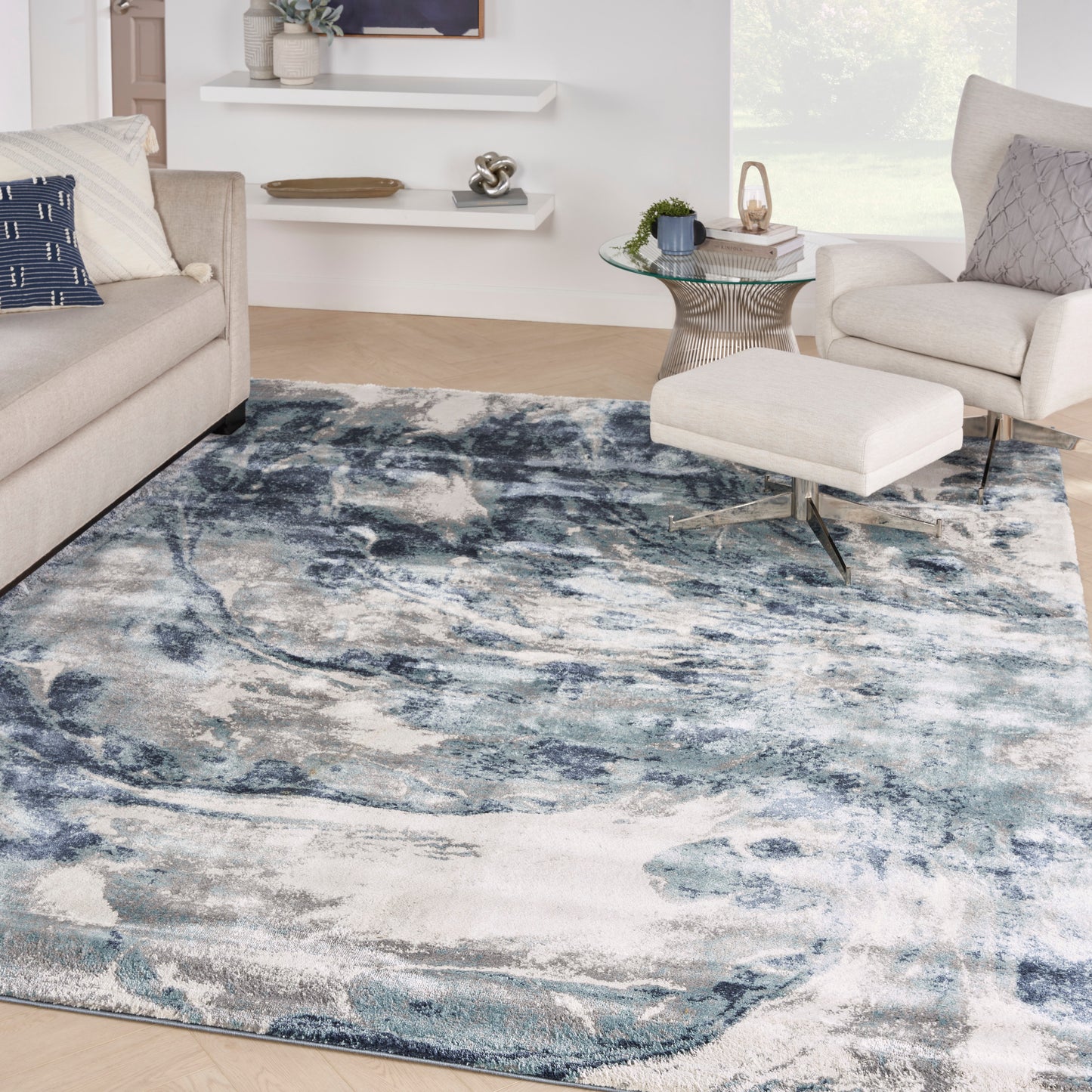 Abstract blue-gray area rug in a modern living room with beige sofas, round glass coffee table, & a light ottoman.
