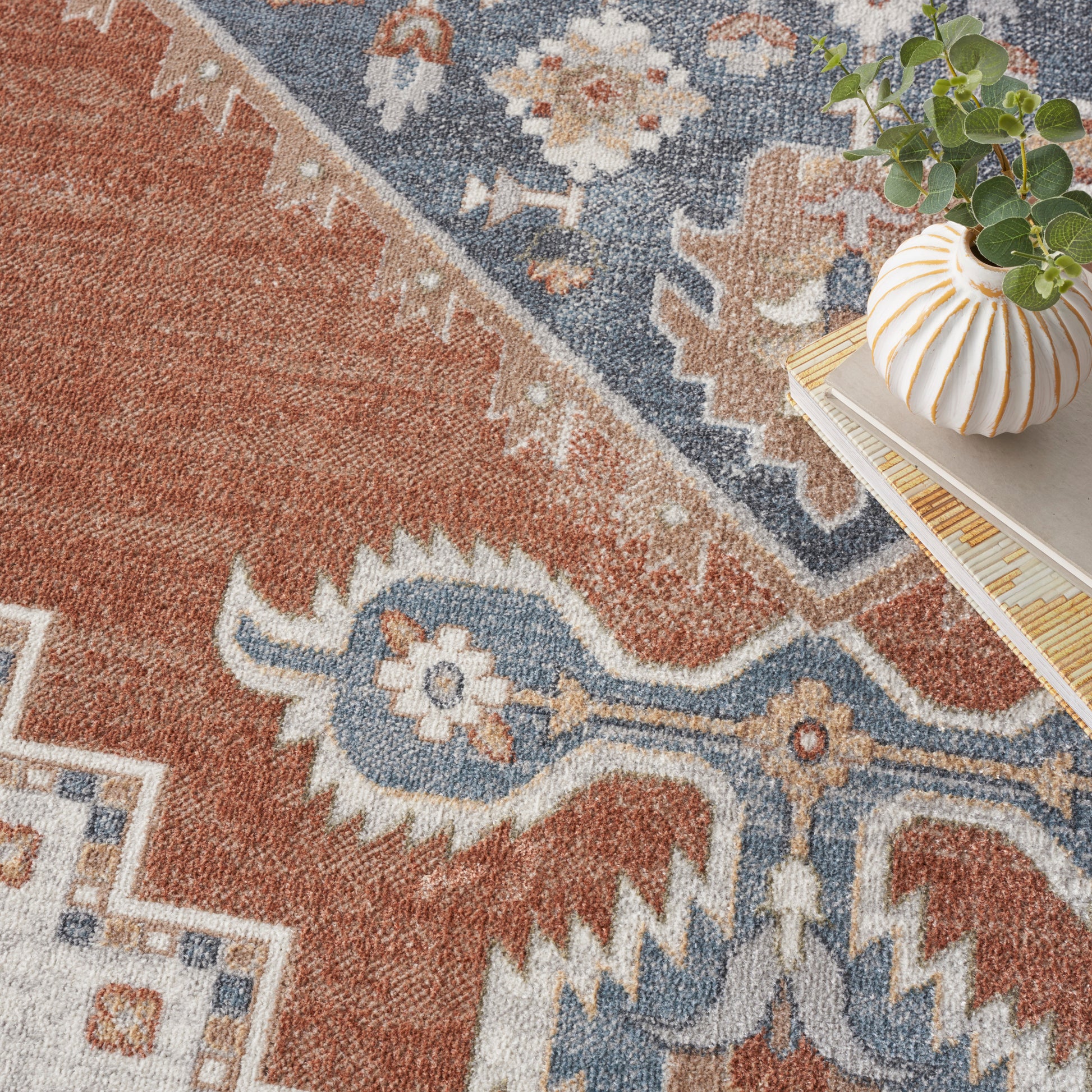 Oriental-inspired area rug in red, blue & cream with floral motifs; ribbed white & gold vase with greenery on stacked books.