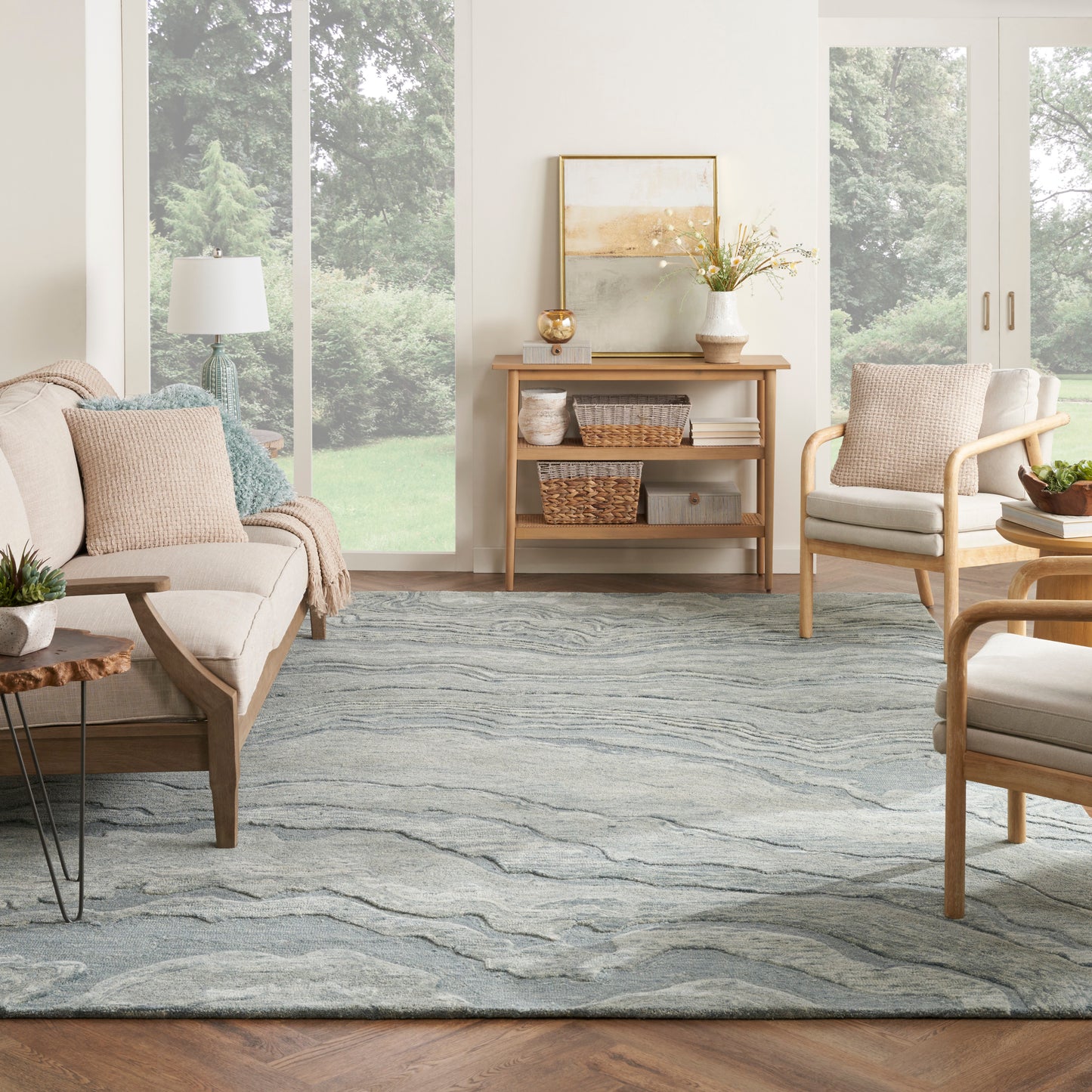 Bright living room with light wood mid-century sofa, lounge chairs, blue-gray area rug, and wicker baskets on a console.