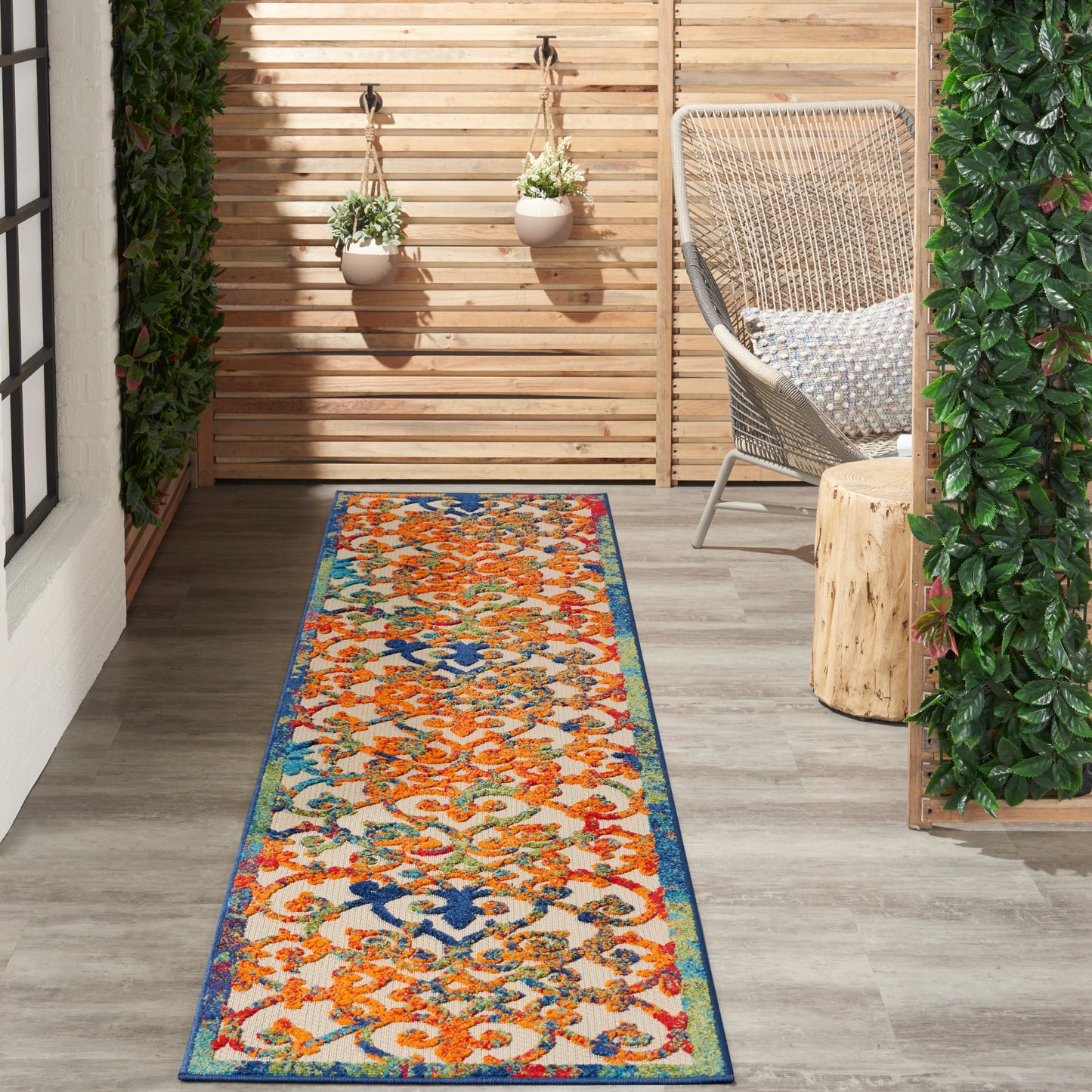 Outdoor patio runner rug with orange floral pattern and blue border on wooden deck beside a wicker chair.