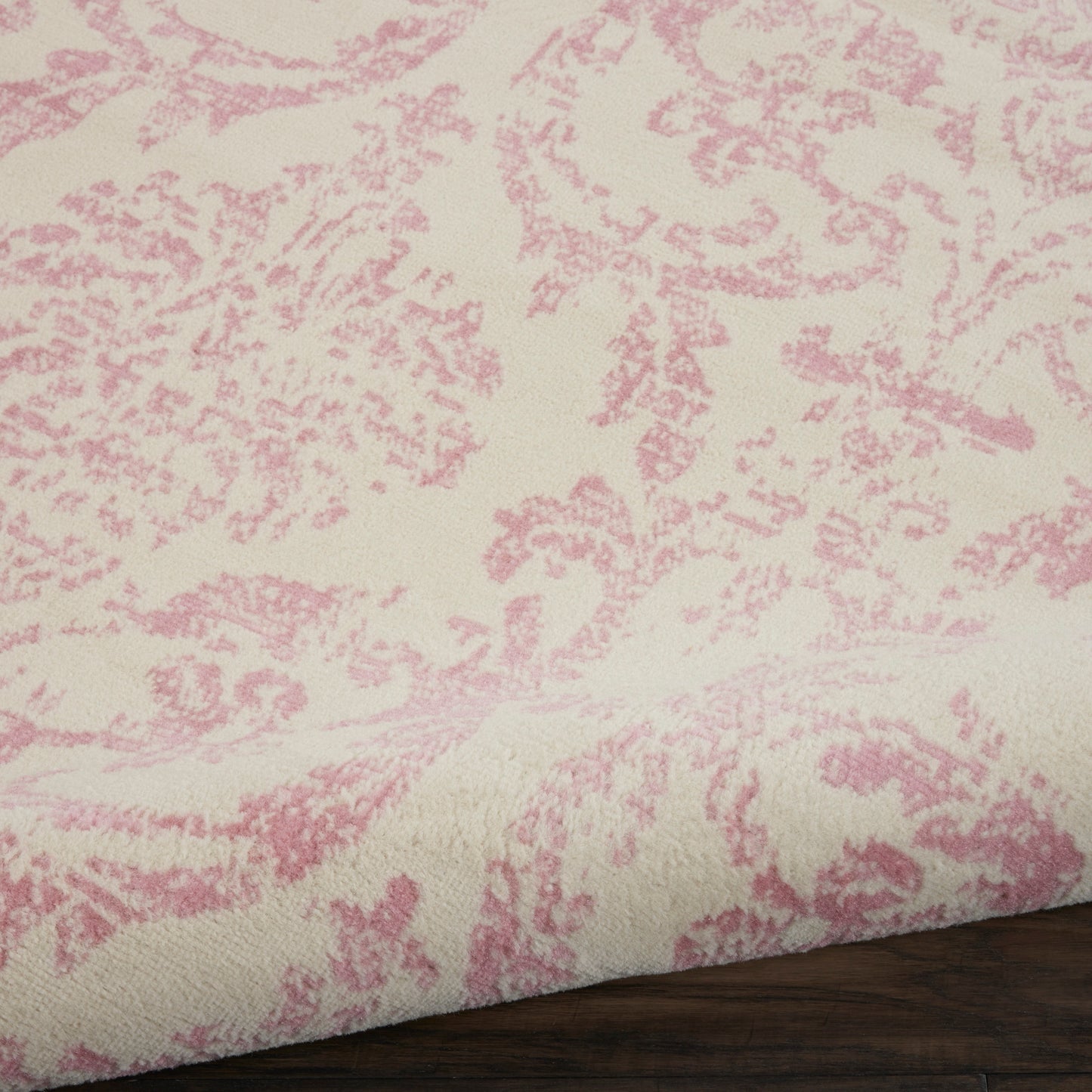 Cream upholstery fabric with pink damask floral pattern, folded edge resting on dark wooden surface.