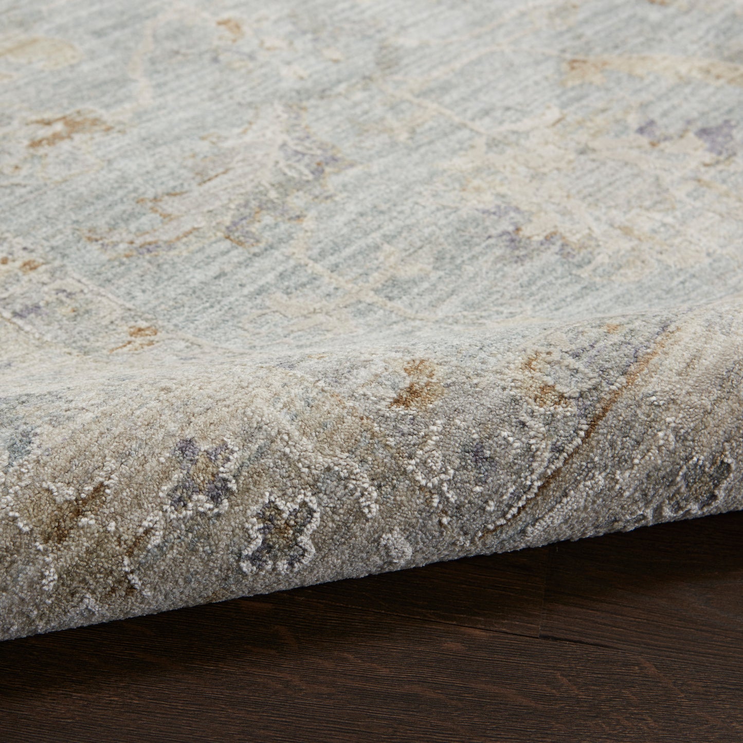 Blue-gray distressed area rug with ivory & tan floral motifs, soft textured pile, edge curling on dark wood floor.