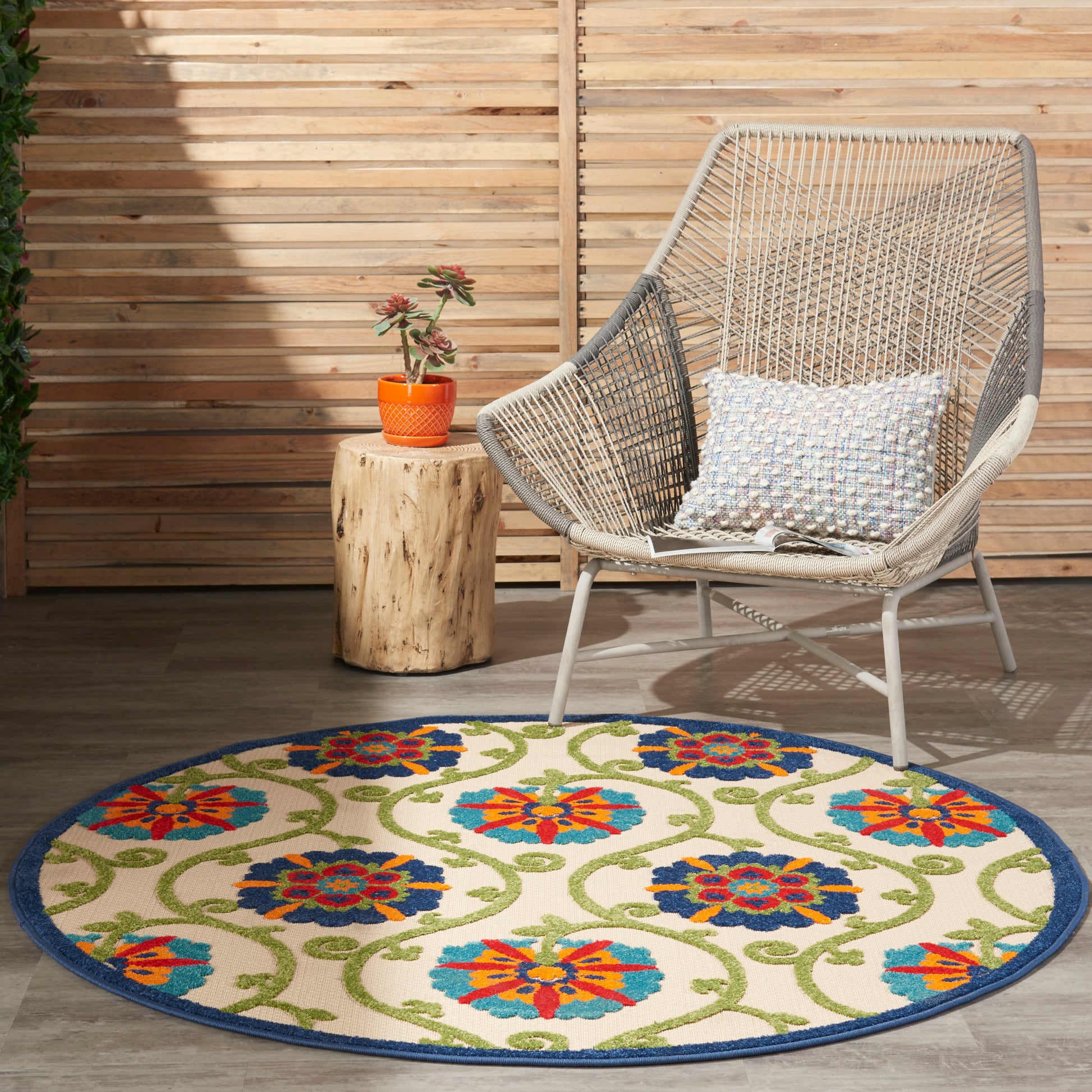 Round floral rug with blue border, beige base and bright orange, blue and green flowers, wicker lounge chair nearby.