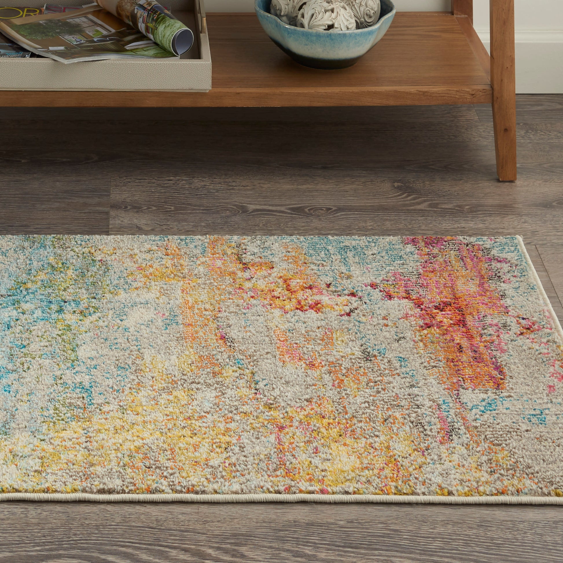 Abstract multicolor area rug with gray base and yellow, blue, pink splashes on dark wood floor.
