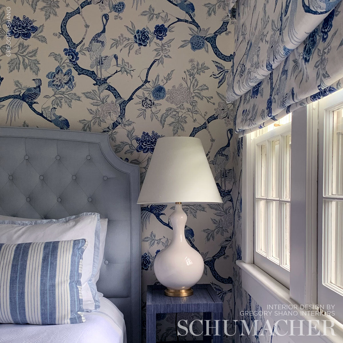 Blue floral wallpapered bedroom with a tufted gray-blue headboard, striped pillow, and white ceramic lamp on a blue nightstand.