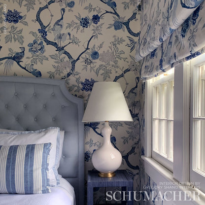 Blue floral wallpapered bedroom with a tufted gray-blue headboard, striped pillow, and white ceramic lamp on a blue nightstand.