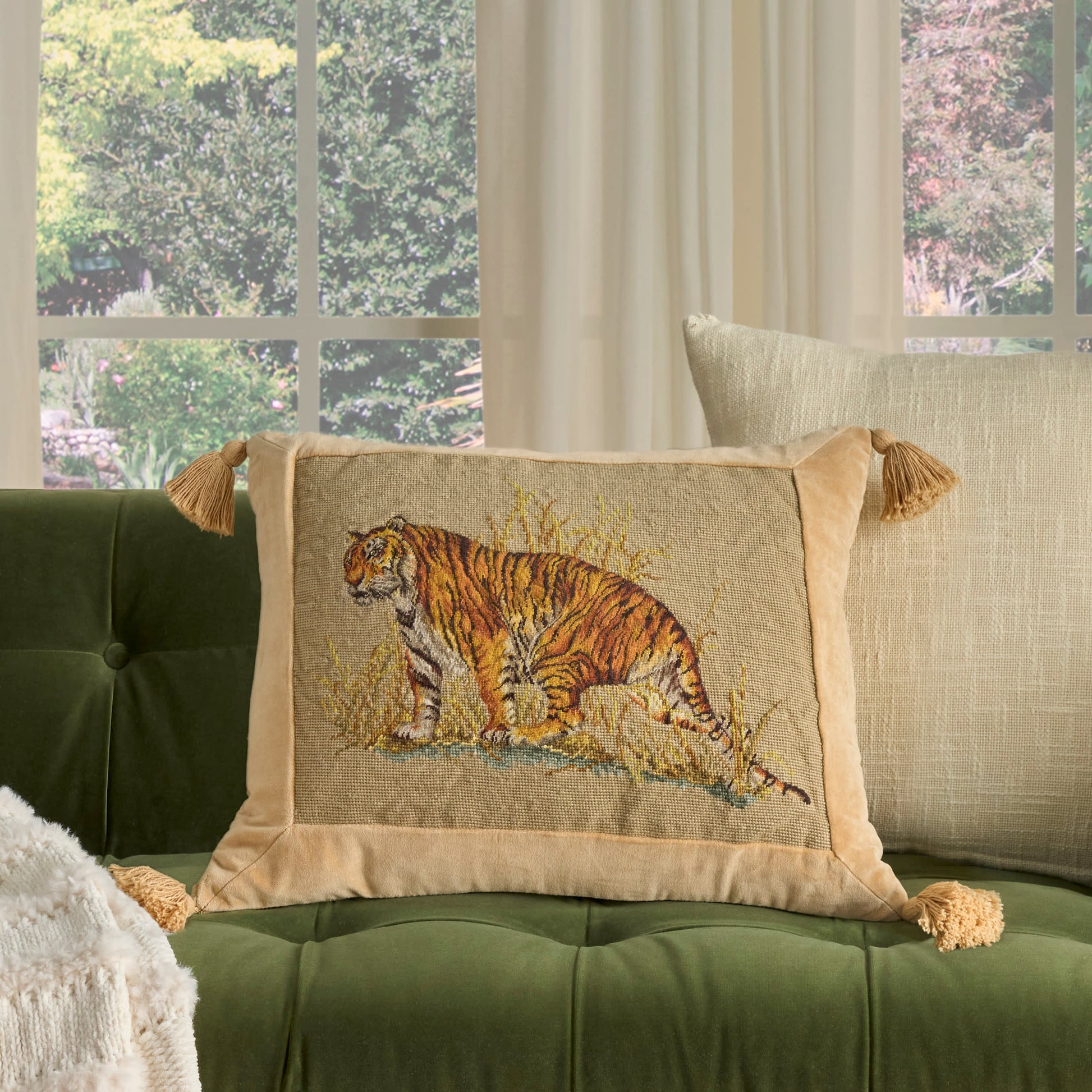 Beige bordered tiger-embroidered decorative pillow with tassels on a green tufted sofa & cream curtains.