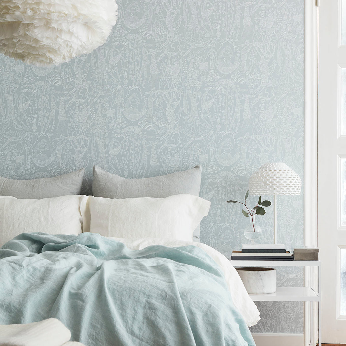 Light blue linen duvet on white bed with gray cushions, pale damask wallpaper, bedside white table, textured lamp and plant.