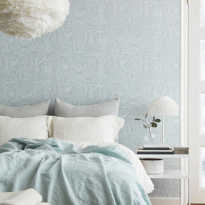 Light blue linen duvet on white bed with gray cushions, pale damask wallpaper, bedside white table, textured lamp and plant.