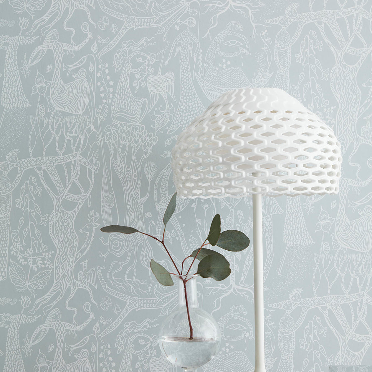 White woven dome lamp on slim stand beside glass vase with red-stem plant and green leaves against blue patterned wallpaper.