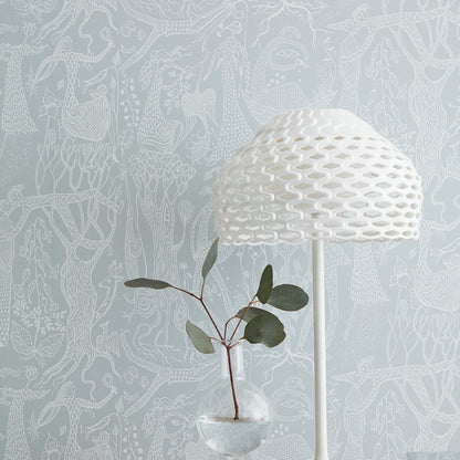 White woven dome lamp on slim stand beside glass vase with red-stem plant and green leaves against blue patterned wallpaper.