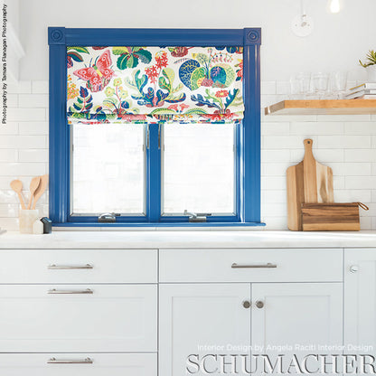 Blue window frame with a tropical butterfly print Roman shade over white subway tile kitchen, white cabinets, and wooden cutting boards.
