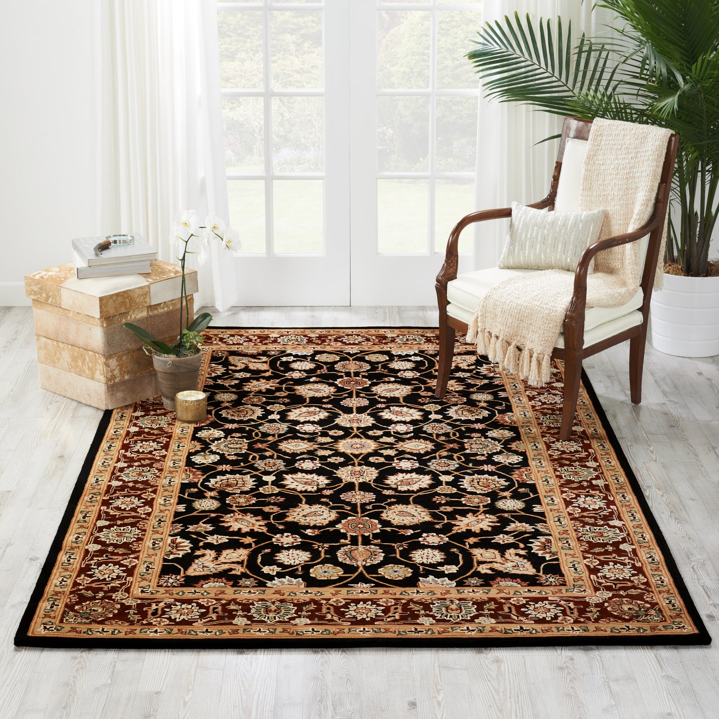 Black oriental area rug with gold and red border placed in a bright living room beside a wooden chair and potted plants.